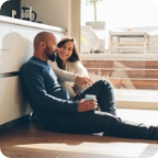 Couple sits on the floor of their kitchen chatting while man holds a cup of coffee.